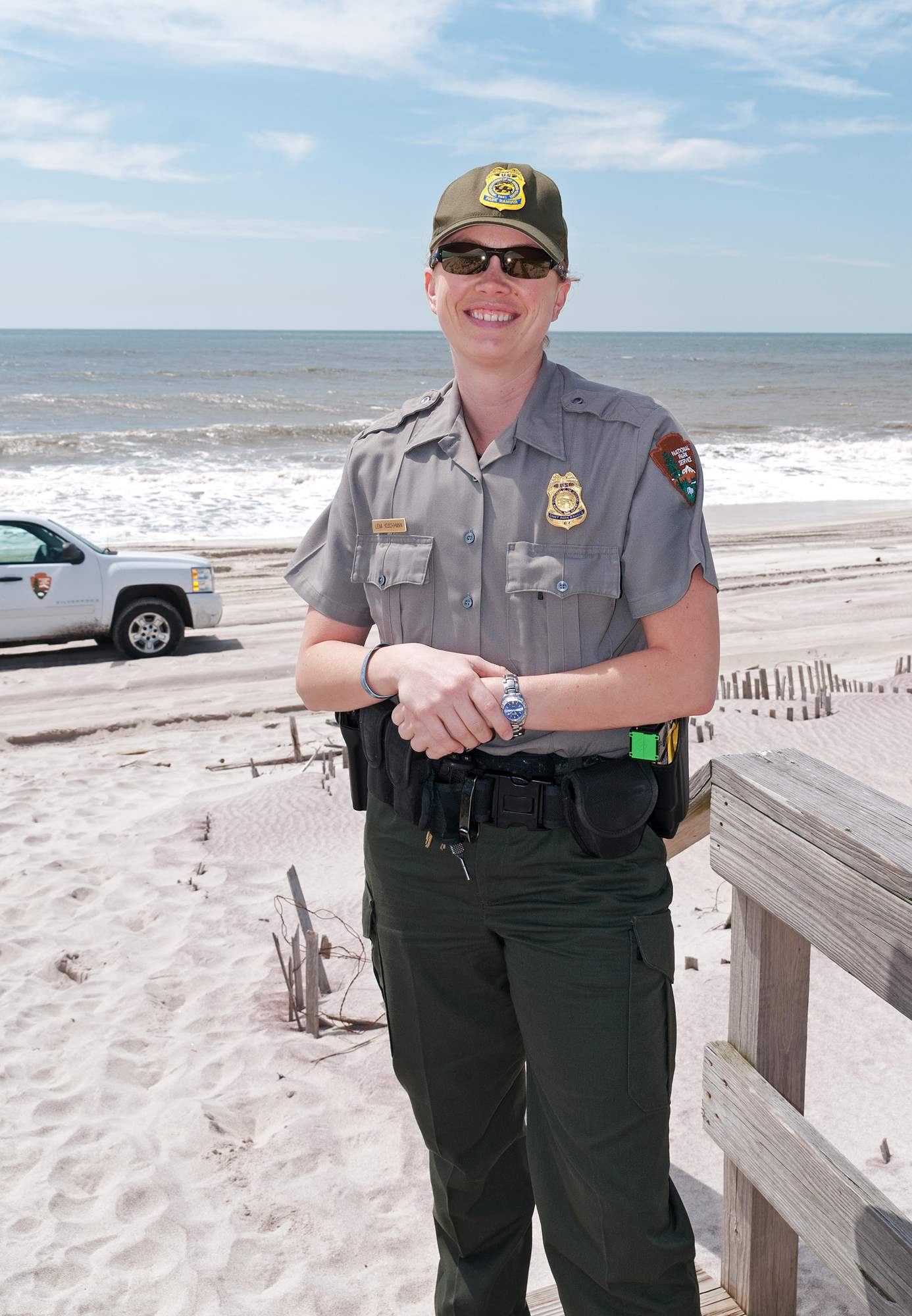 Lena Koschmann stands on a beach wearing NPS uniform and ball cap. She has a shield-shaped badge with an eagle perched on top pinned to her shirt.